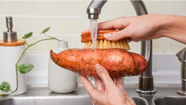 Hands scrubbing a sweet potato in kitchen sink.